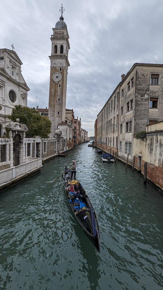 Looking down a canal, a gondolier pilots his gondola with a couple passengers. In the background a pale stone tower stands with a distinct lean towards the water.
