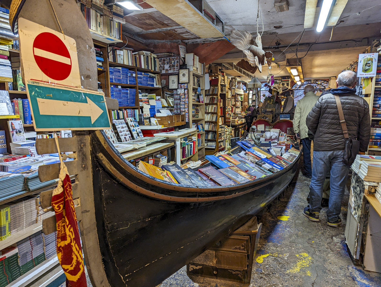 A gondola in the aisle of a bookstore, with books displayed in it. On each side are shelves with other books. Francesco is walking away from the camera.