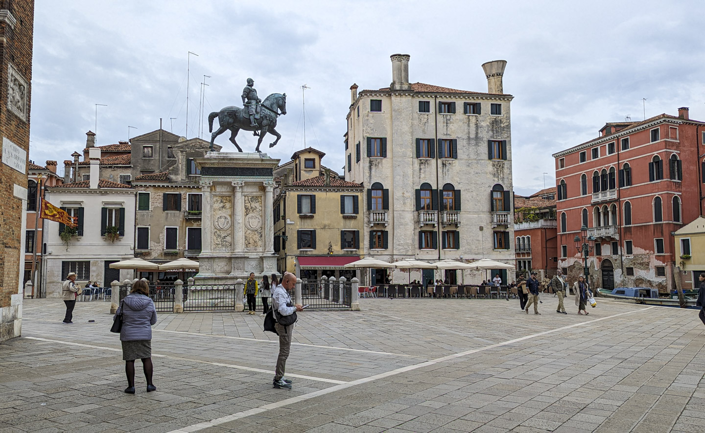 A plaza in Venice with a weathered bronze statue, on the top of very large pedestal, of a horse with rider. The horse looks like it is about to take a step. One of the canals can be seen on the right of the photograph. A venetian flag is flying on the left edge of the photo.