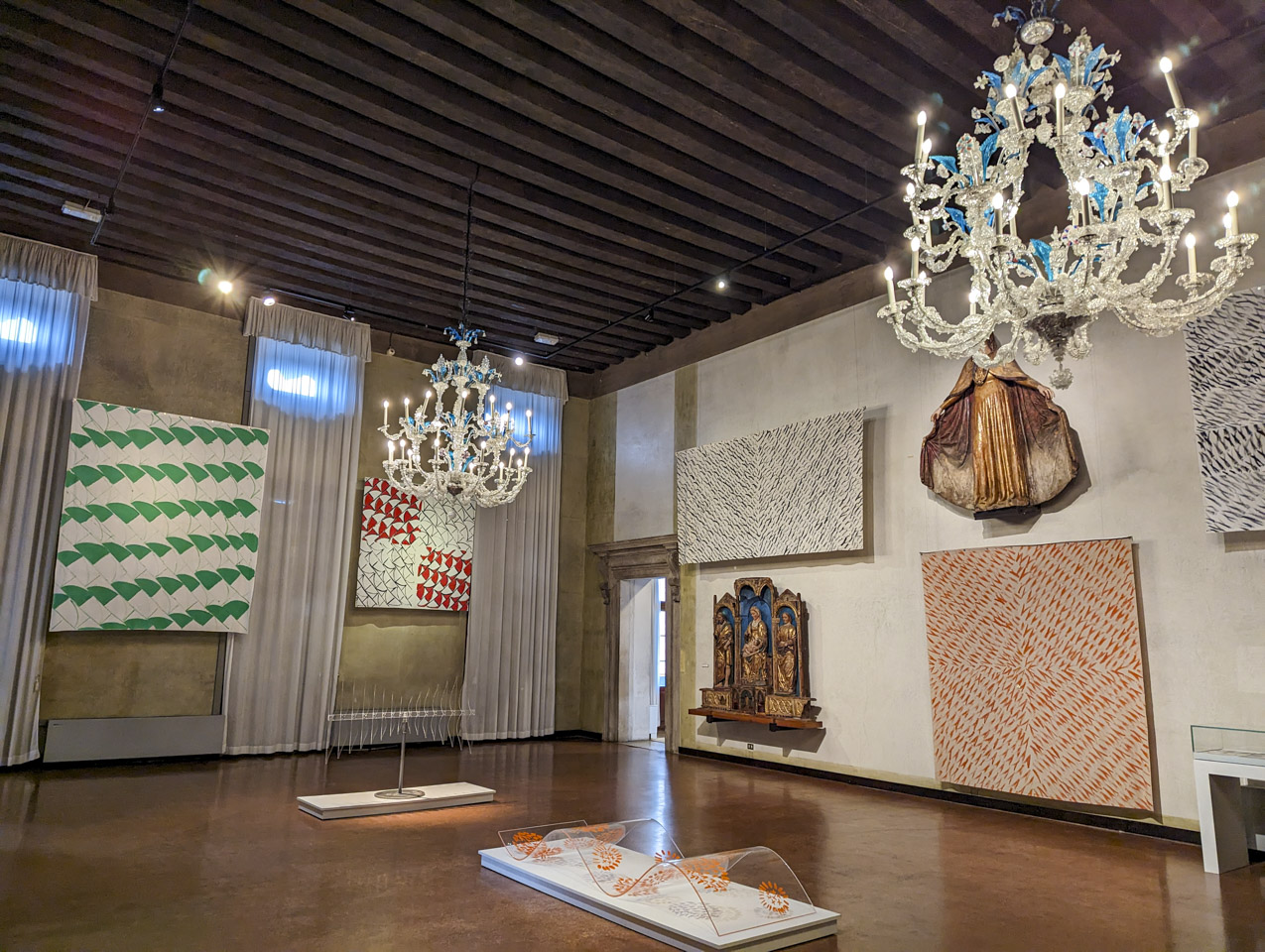 A museum room with many geometric paintings on canvas as well as some glass structures on the floor.