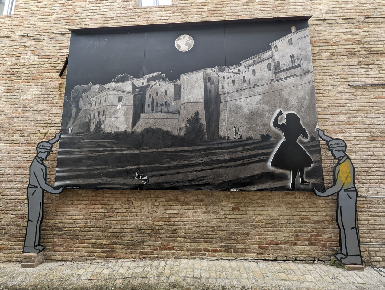 A large print against a stone wall. The largest part of the print medieval village. The foreground has a sillhouette that looks like Alice in Wonderland. In the sky is the moon, but it shows the continents of Earth. A worker is painting the city walls.  There is a cat on the ground. In the background is a nun in her habit standing next to an alien.