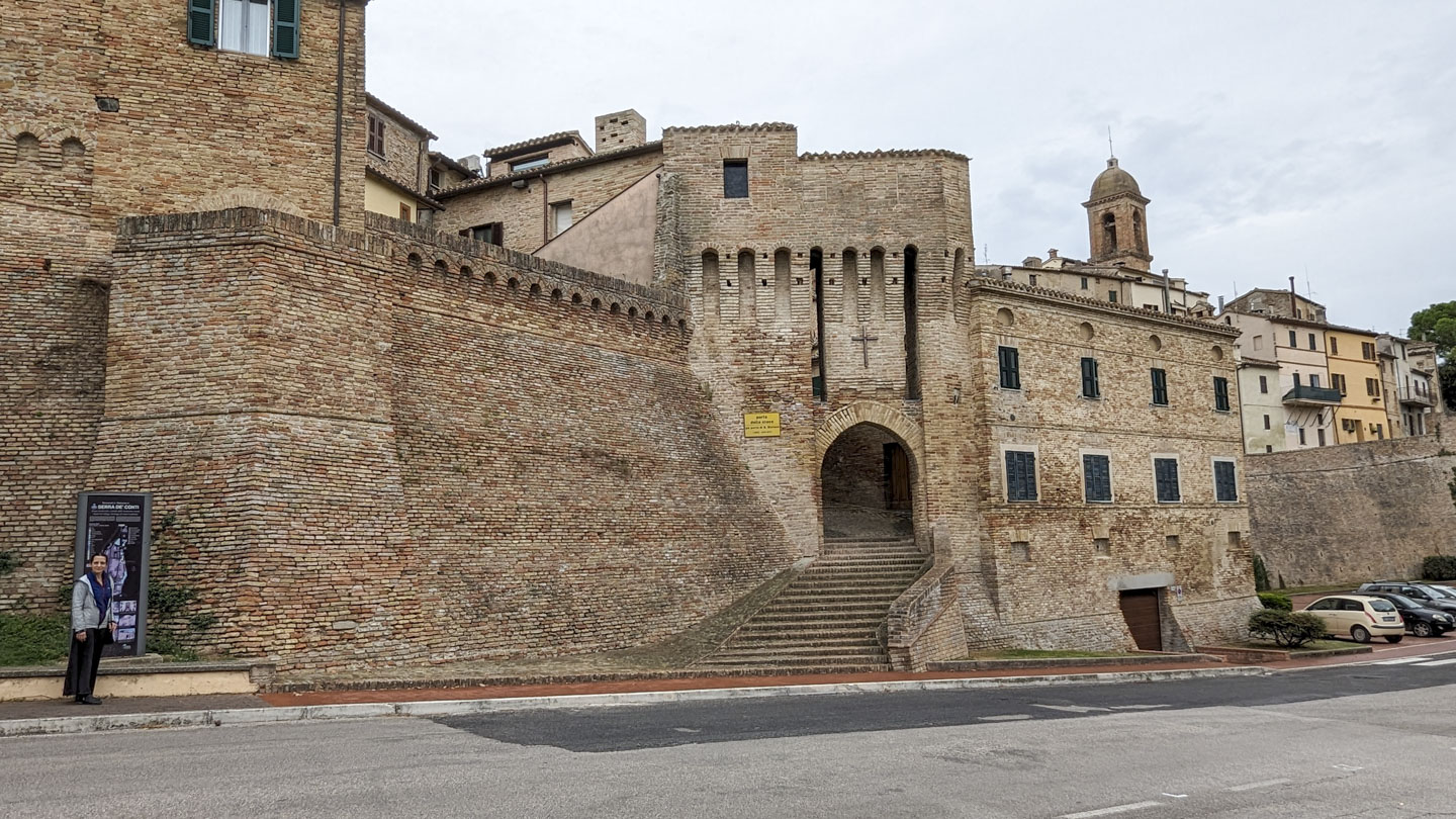 Anne is standing in front of a sign on the left of the photo, with the large stone walls of the city behind her. In the middle is an arched entrance with a cross above it, and there is a tower in the background.