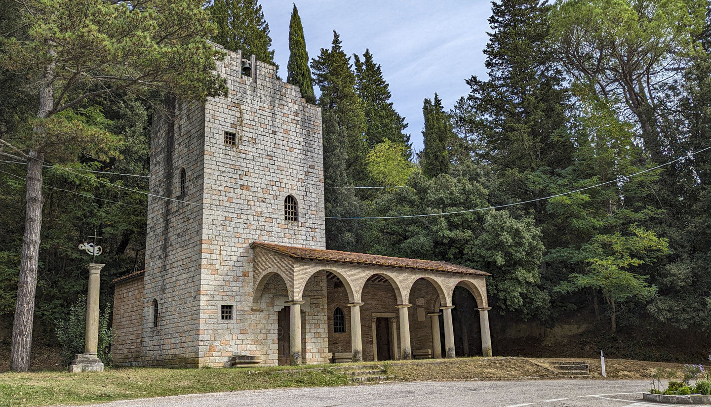 Stone church with a bell tower on the left, that has a very small bell on the top, and an arched covering along the front of the building.