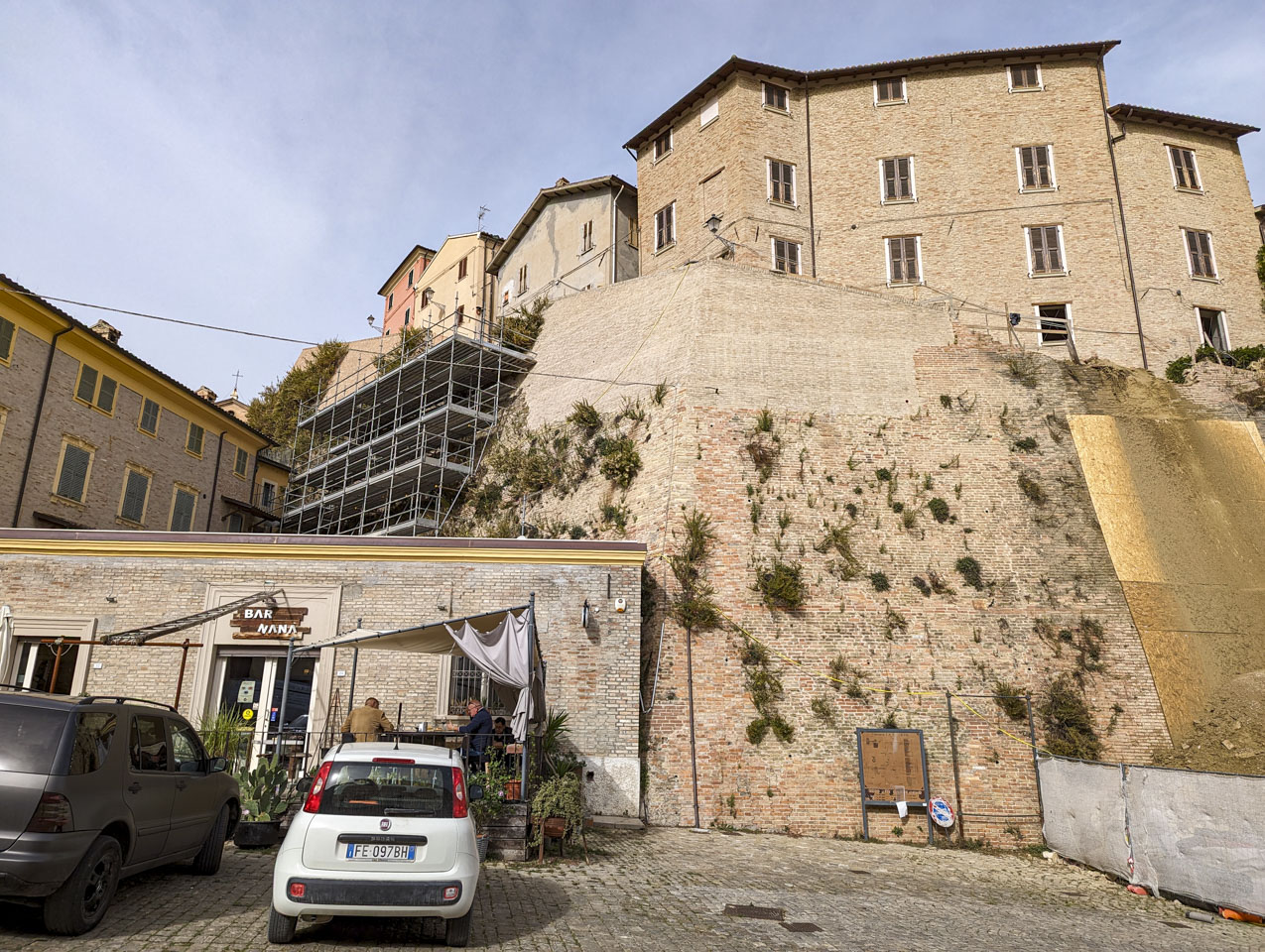 On the left of the photo is Bar Nana with a couple men sitting in front, and behind that is scaffolding going up the old city wall. To the right is a section of wall with the top portion being new and the bottom portion being older with plants growing out of spots.