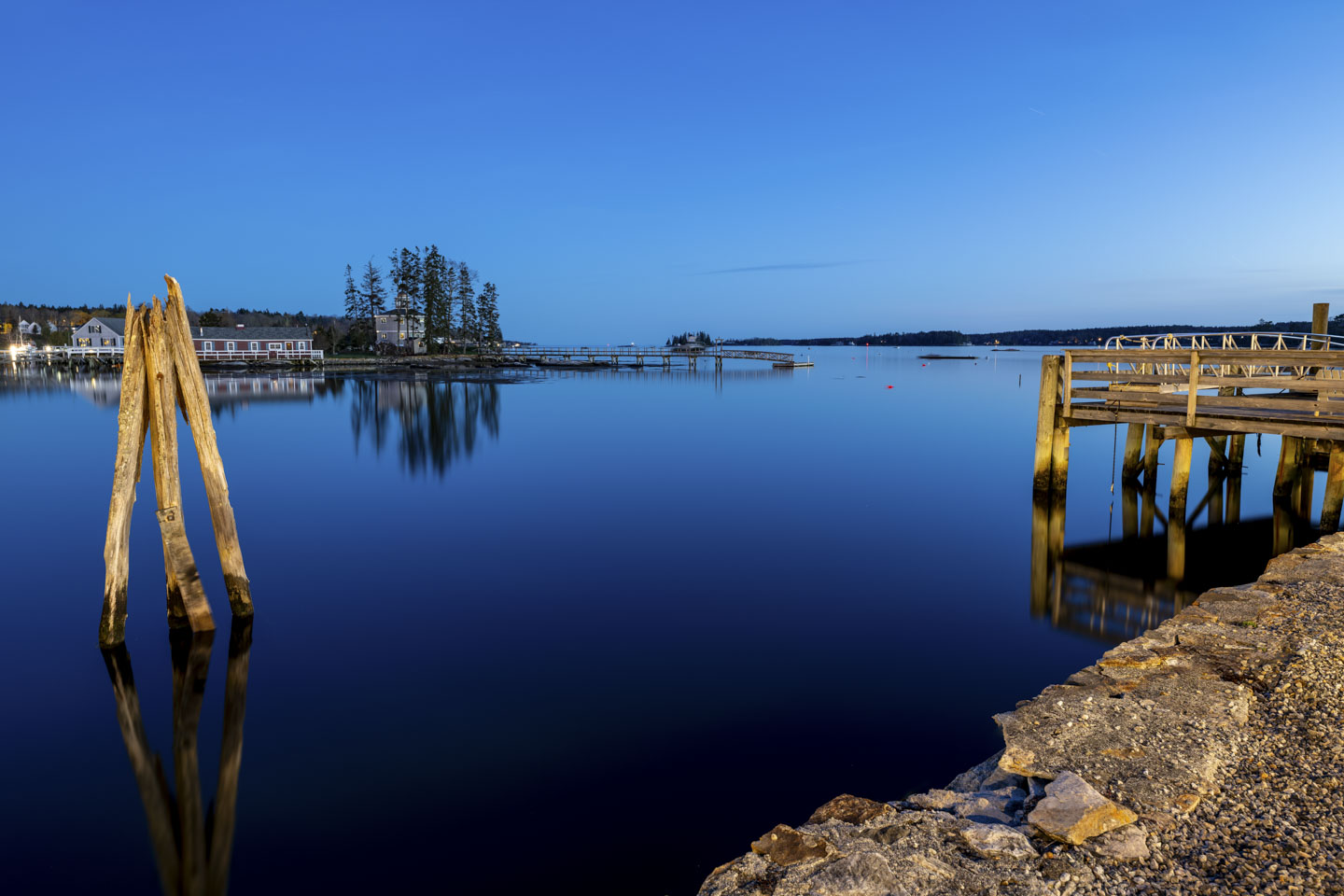A view of Boothbay Harbor in the evening, with some old pilings on the left and a pier on the right. A long pier can be seen in the distance.