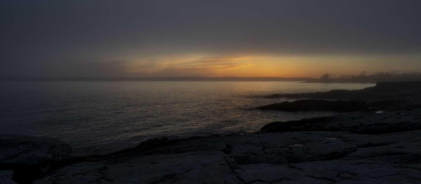 The sun has just set, and fog is moving in, obscuring the view of rocks, water, and trees.