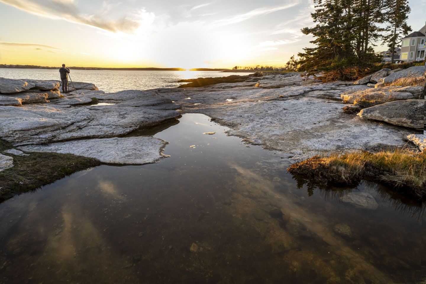Paul is standing on rocks on the left, with his tripod. Rocks are also on the right. There is a large tidepool in the front, and then more open water. The sun is setting over land on the opposite shore.