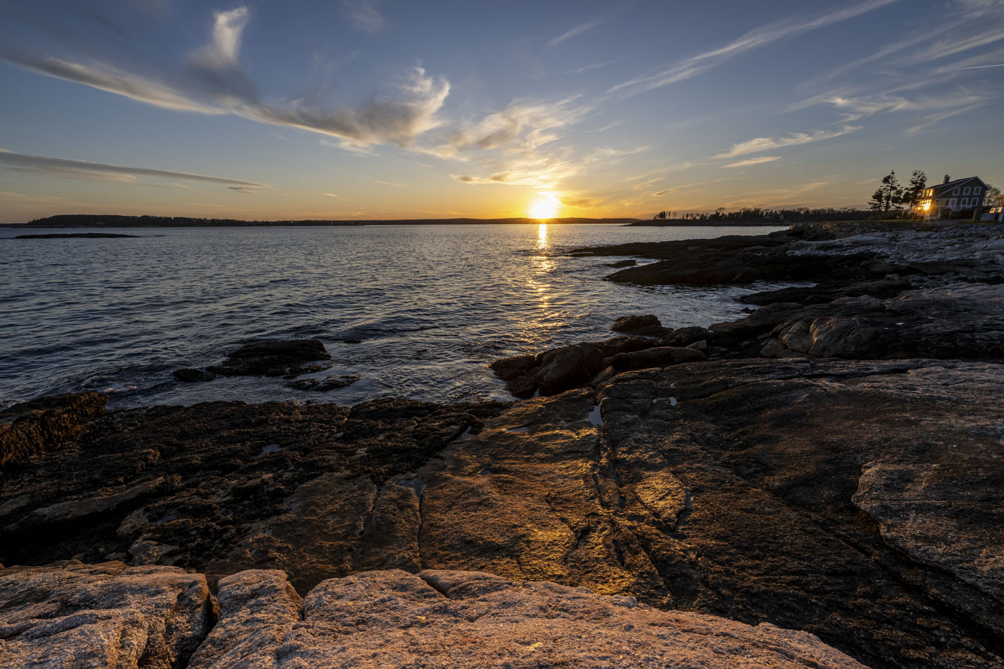 The low sun reflects off the window of a house on the right of the picture, with rocks and water making up most of the photo.