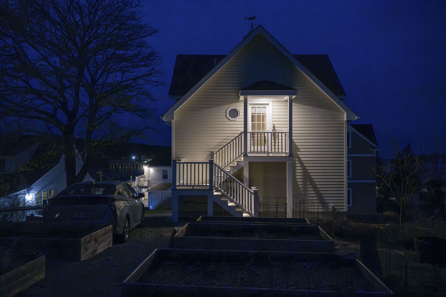 A tan building with a door on the second floor that has a landing and stairway. The light is on outside of the door. The sky is dark blue, and a tree is on the left.