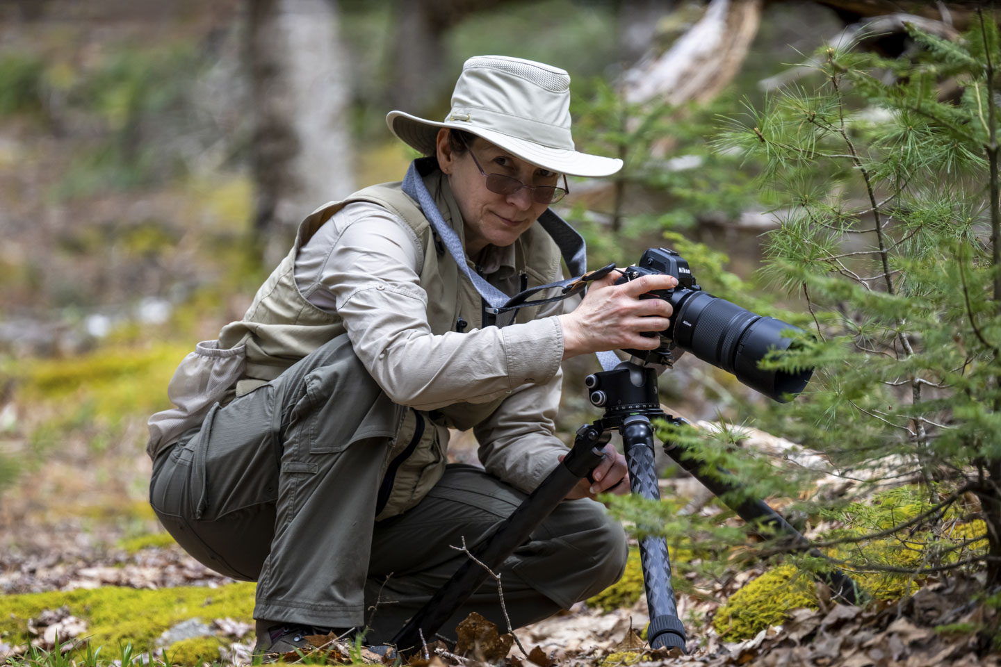 Anne is squatting near the ground, wearing a brimmed hat, while holding her camera that is on a tripod.