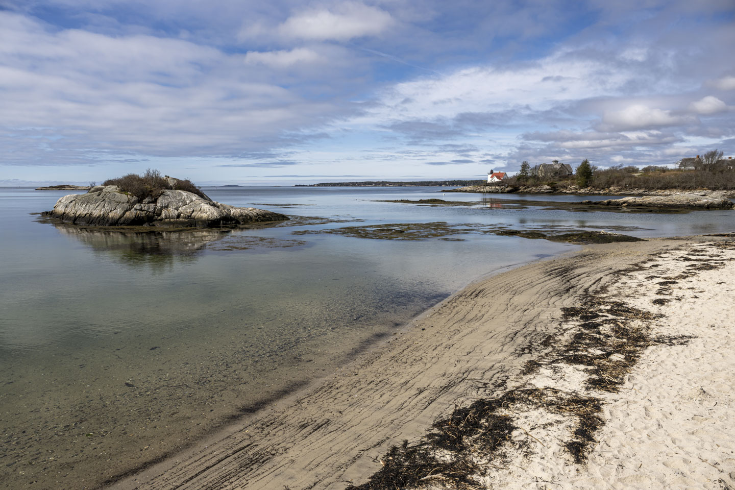 A beach is angling away and to the right. There is a tiny rocky island sticking out from the shallow water. In the distance on the right can be seen a lighthouse.