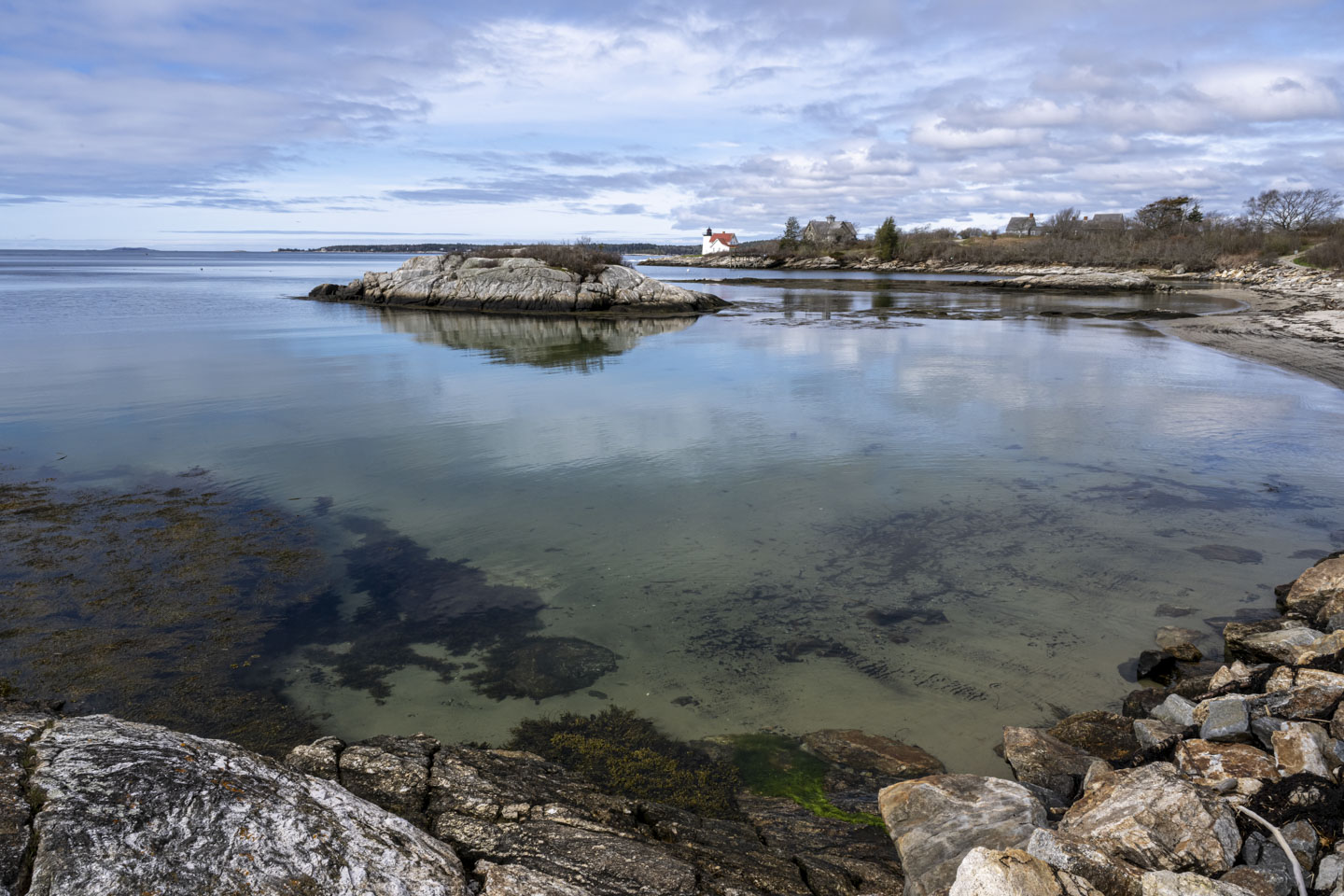 Shallow water wraps around a small island, and in the distance a white lighthouse with a red roofed house is on a point of land.