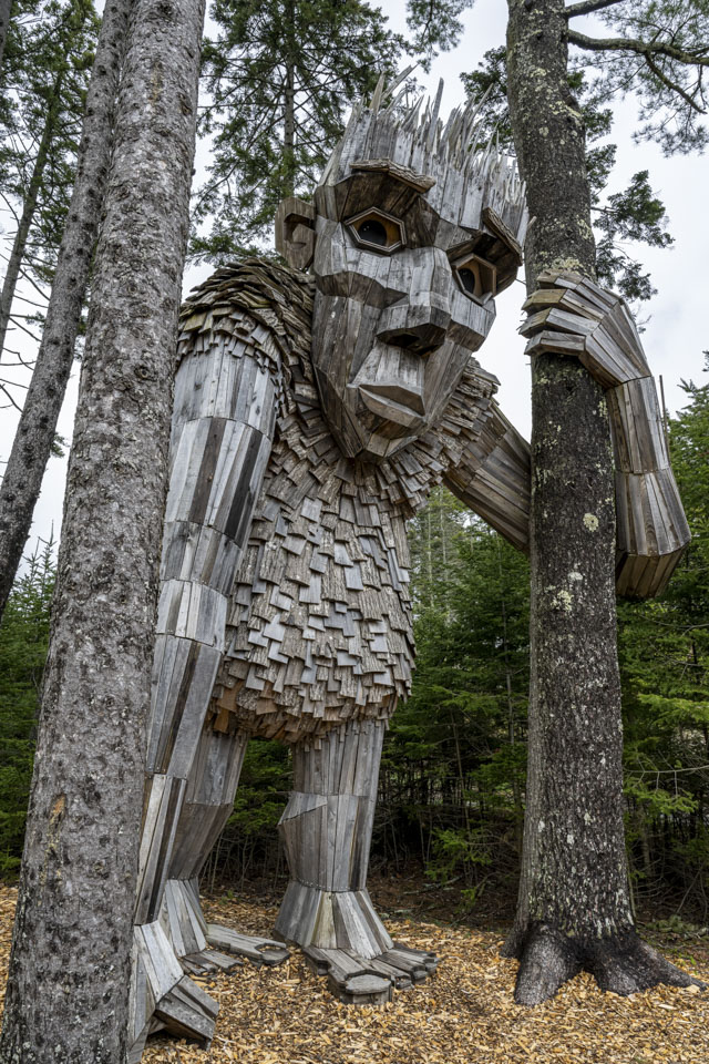 A large wooden troll looks down on use while holding a tree.