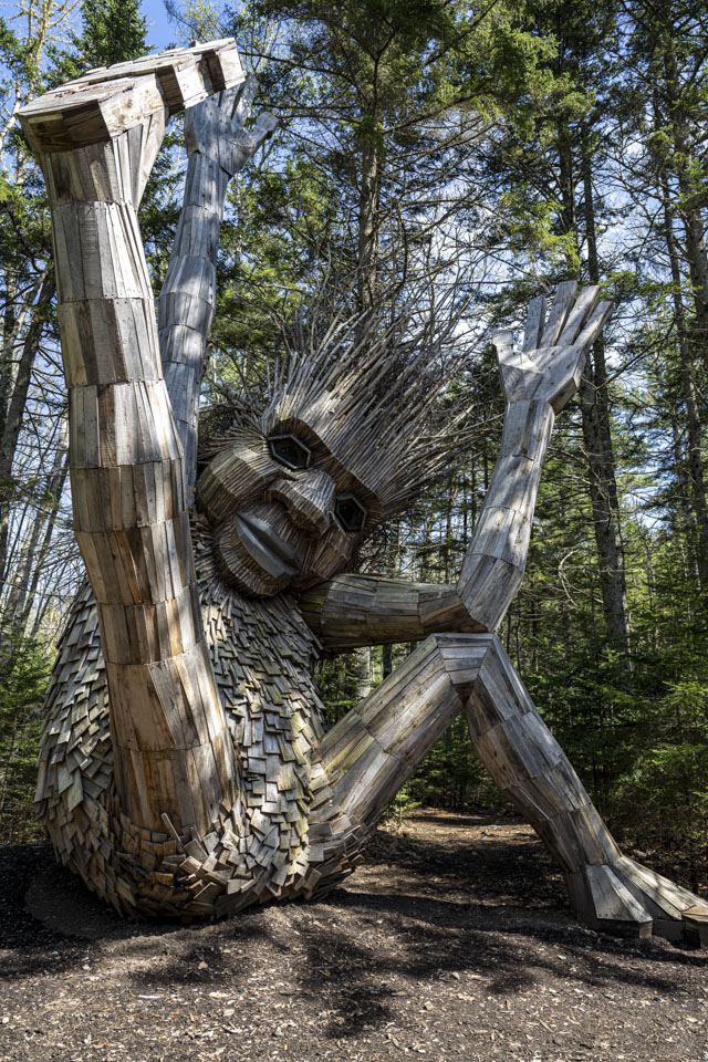 A wooden troll with spiky branches for hair seems to be having fun, having kicked up a leg and its arms.