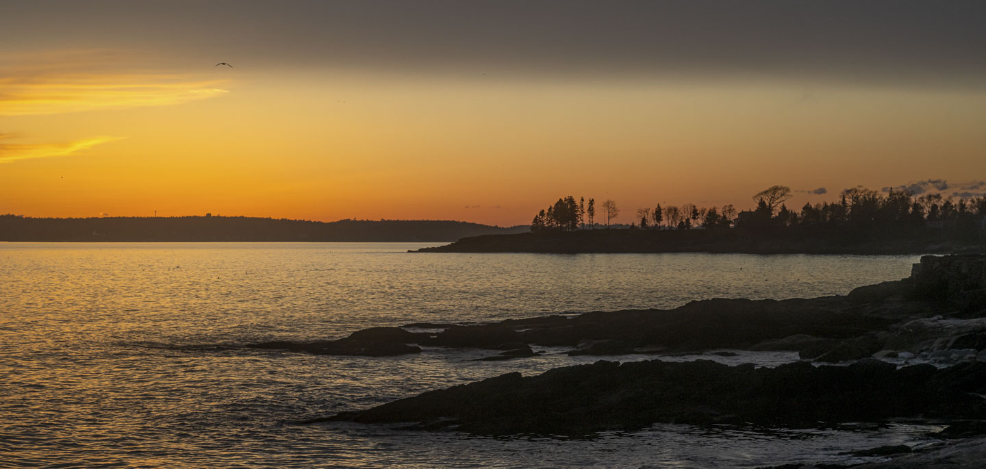 After sunset, with an orange glow on the horizon. Rocks, waters, and the outline of trees can be seen.