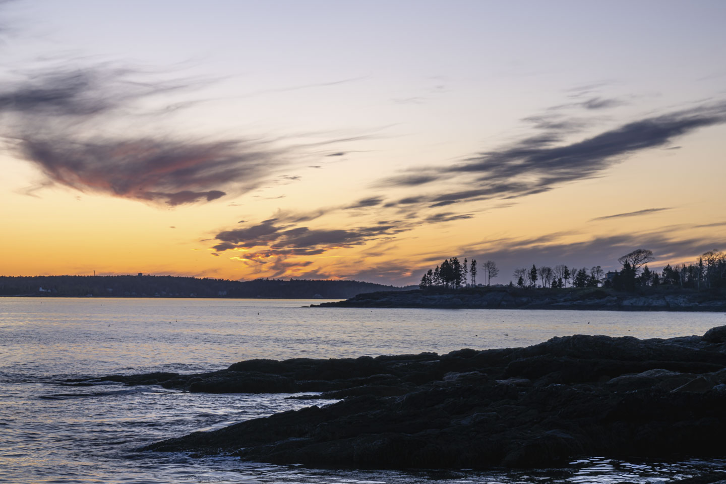 After sunset, with an orange glow on the horizon. Rocks, waters, and the outline of trees can be seen.