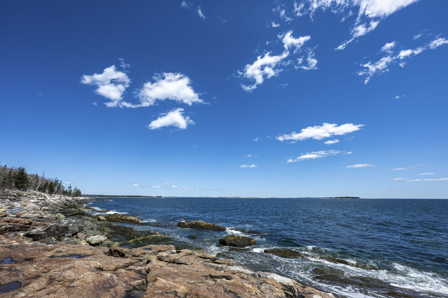 Blue sky with some puffy white clouds is above a rocky coast with blue water.