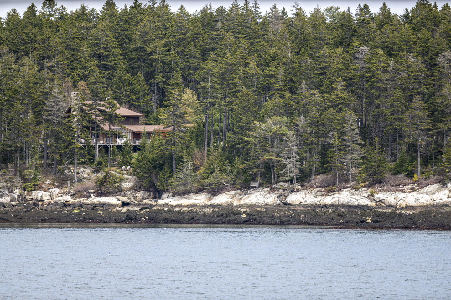 Looking at a rocky coast on the other side of river water, a small bench can be seen on the edge of a woods. On the left is a large wood house.