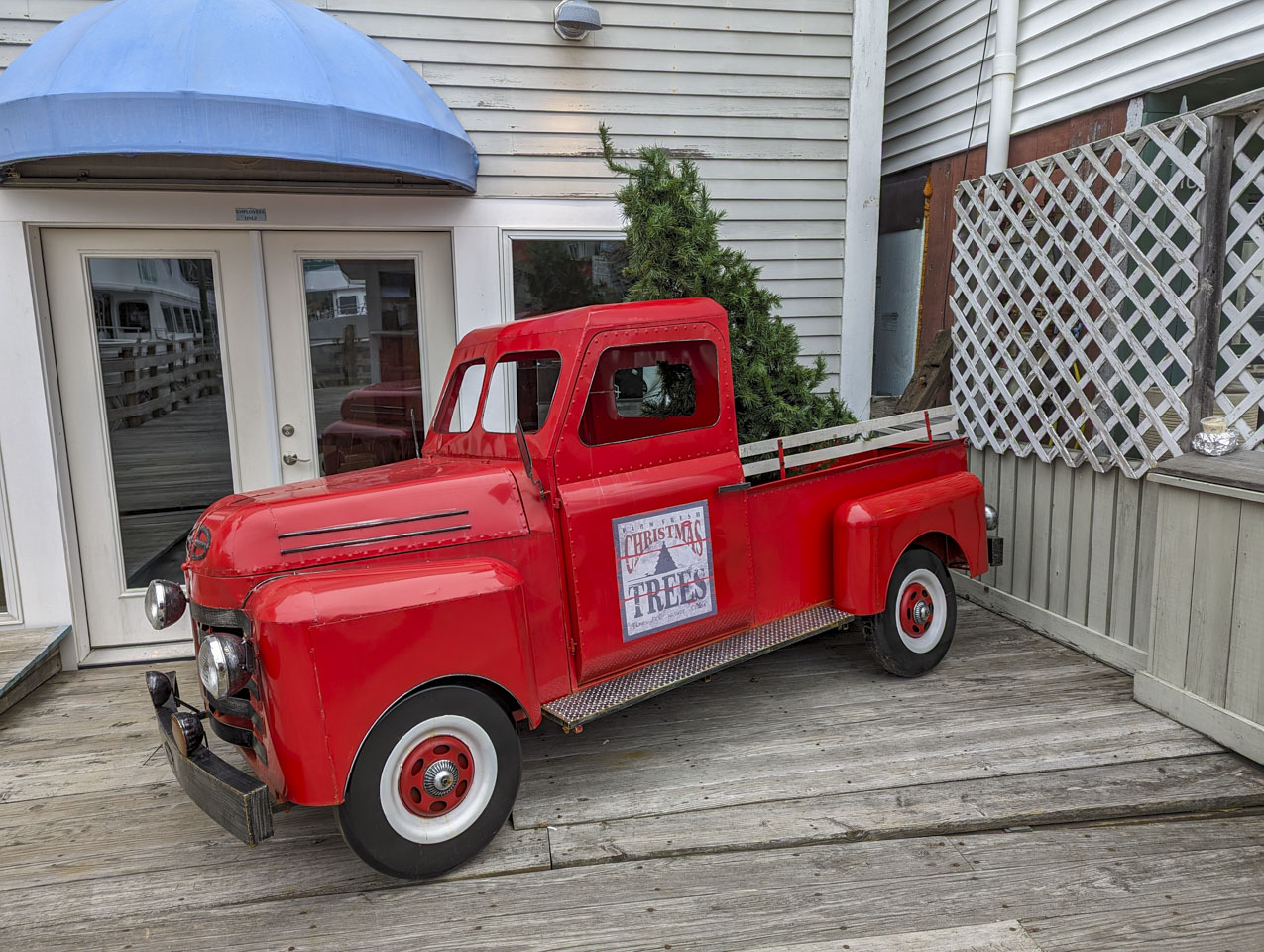 A classic looking red pickup truck with an artificial undecorated Christmas tree in the bed area. It is sitting on wood plants in front what looks like an empty store front.