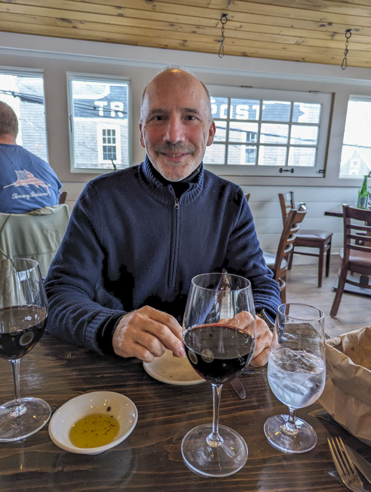 Paul is sitting at a restaurant table. On the table are 2 glasses of wine, a glass of water, and some oil for dipping bread.