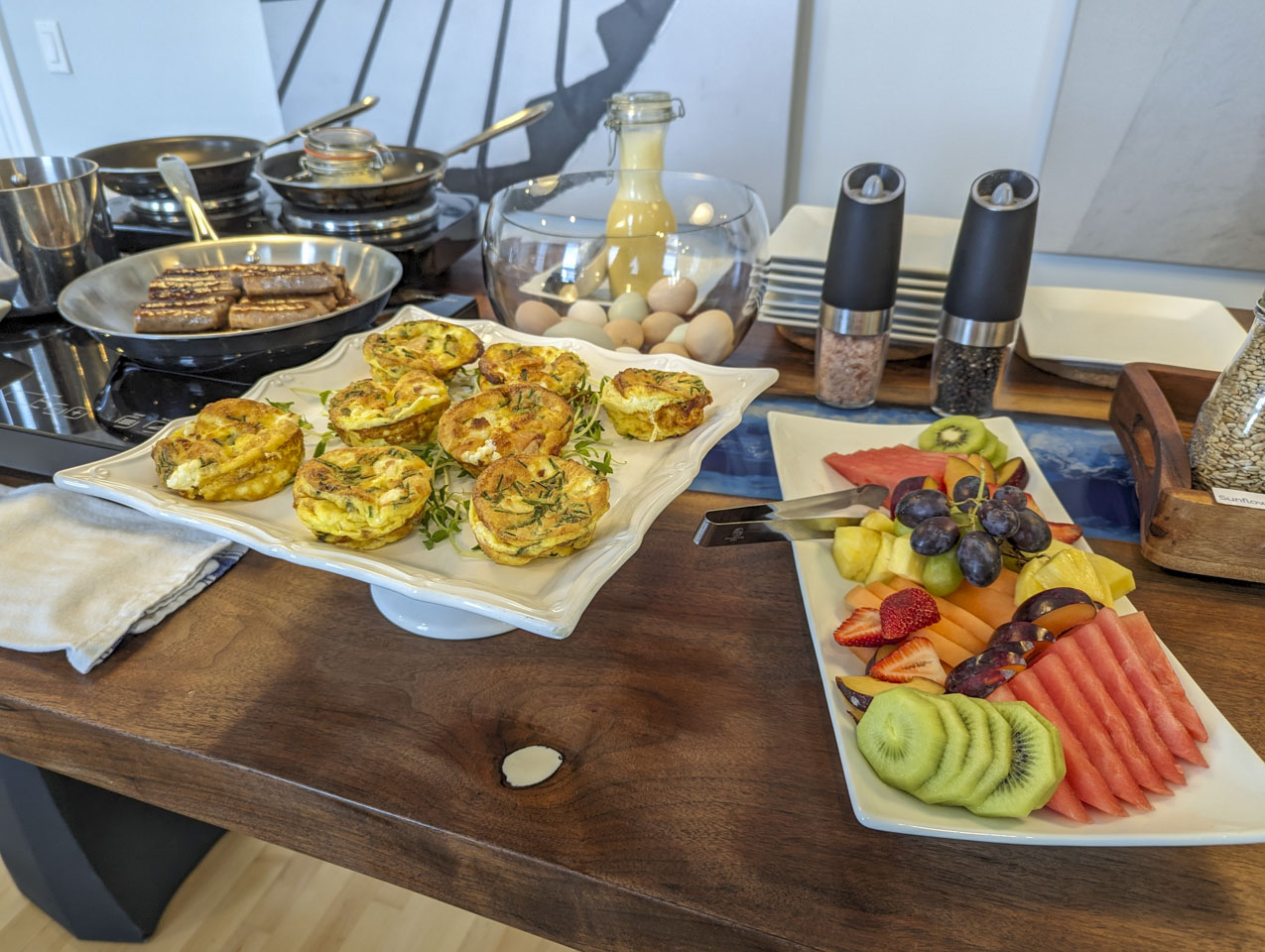 A close-up of the table after more food has been put on it. There is fruit, little souffles, sausage, and whole eggs in a glass bowl.