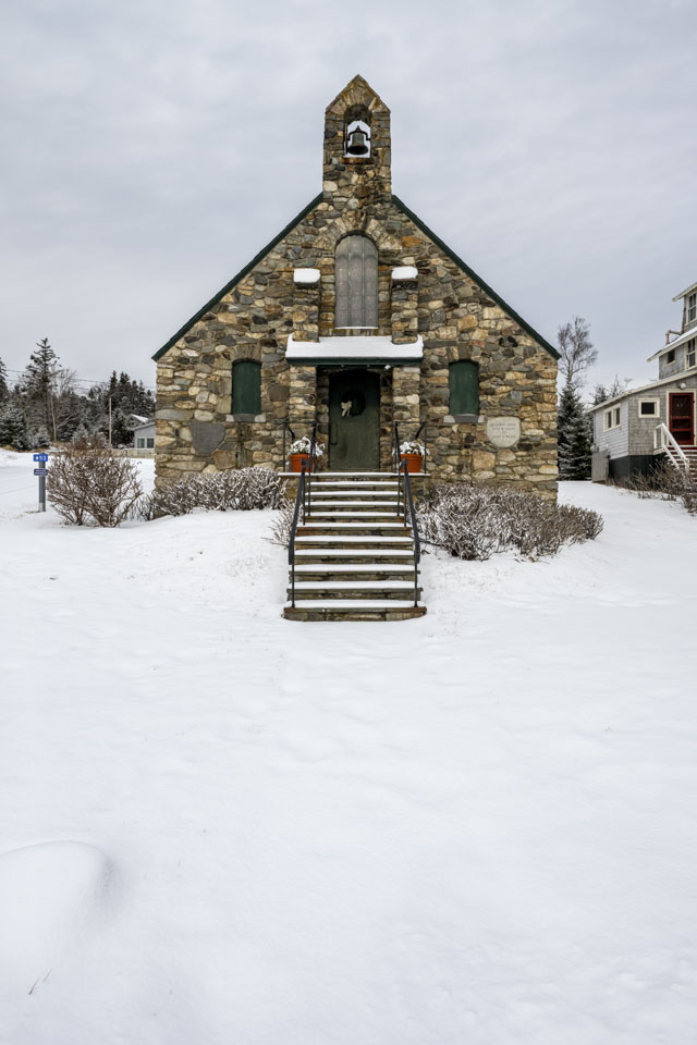 A stone church, with a bell in the tower, has snow-covered steps leading down to a snow-covered walkway.