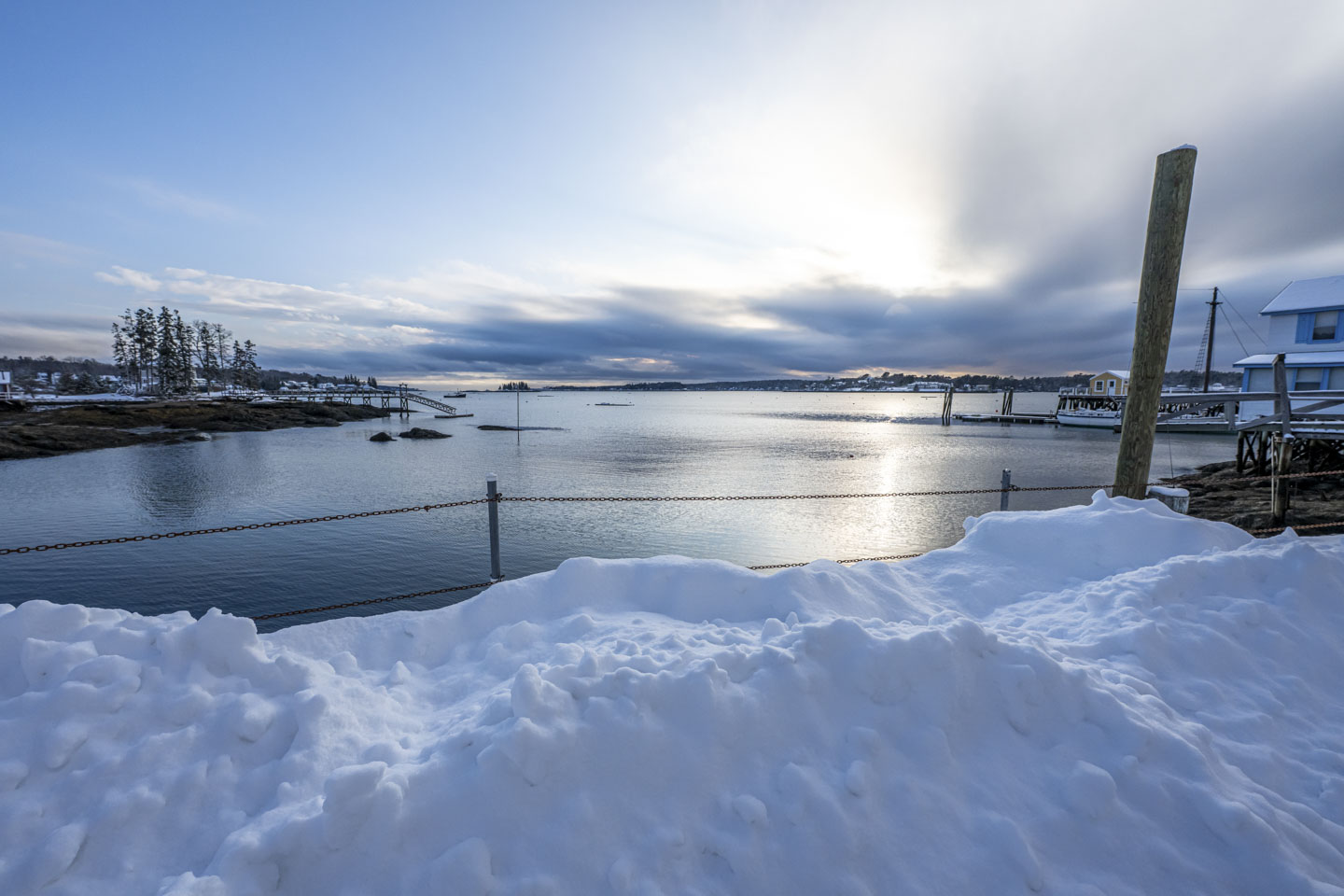 The sun creates a glow behind some thick horizon clouds and gives the water a glow. This is seen by looking past a large mound of snow.