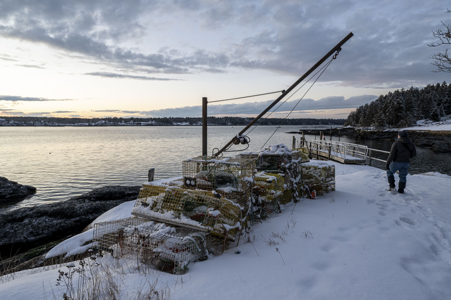 A snow covered dock has snow covered lobster pots on it. Paul is walking through the snow towards the pier.