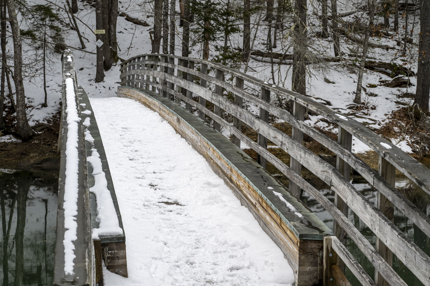A gently arching wood pedestrian bridge is covered with a bit of snow, and a sign that says Vista can be seen on the far side.