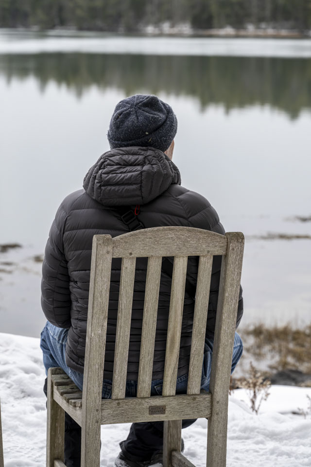 Paul is sitting on a wooden chair in the snow. His back is to us and he is looking to the right.