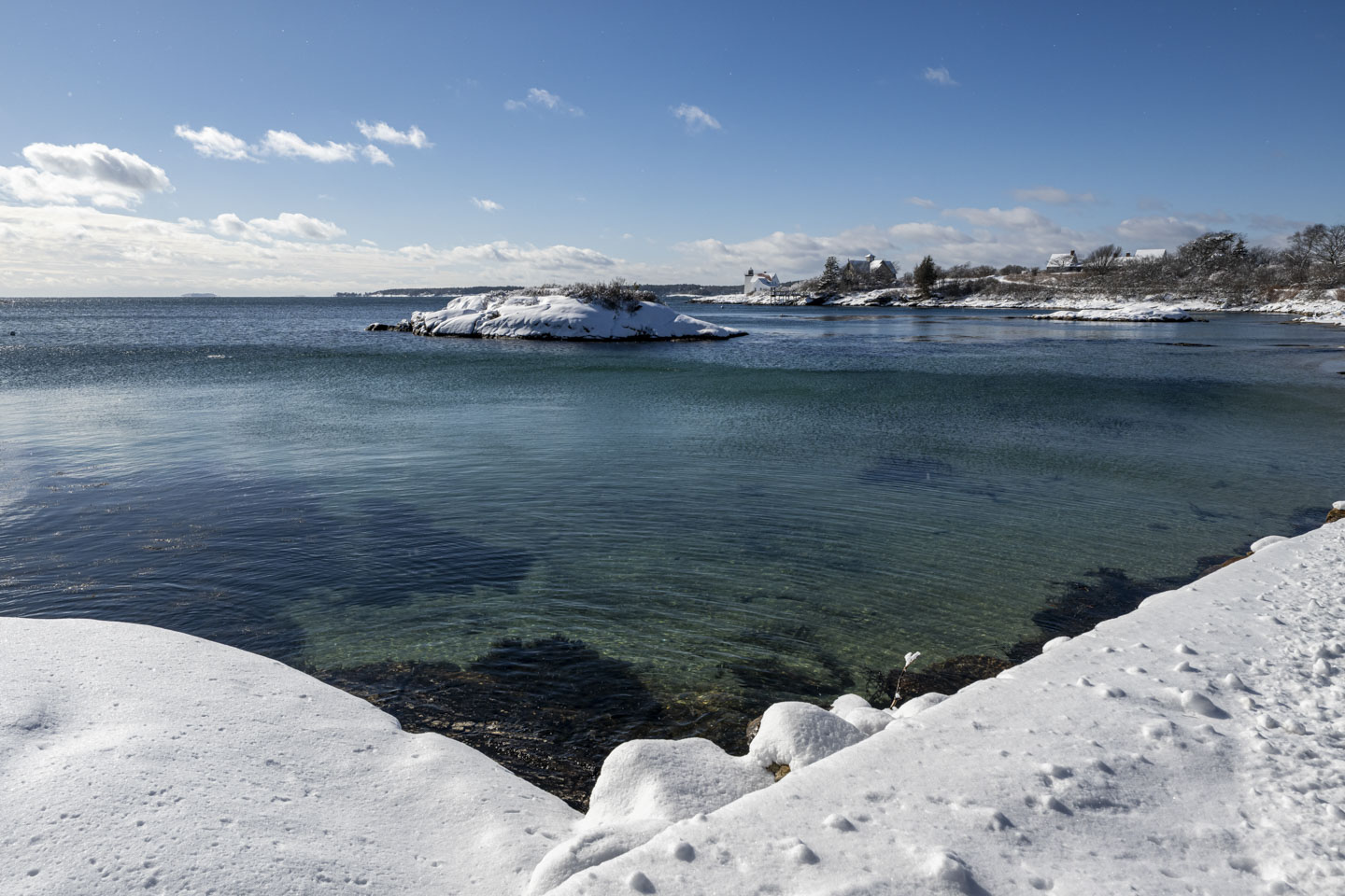 Looking from the edge of a harbor, you can see a snow-covered island and a lighthouse in the distance.