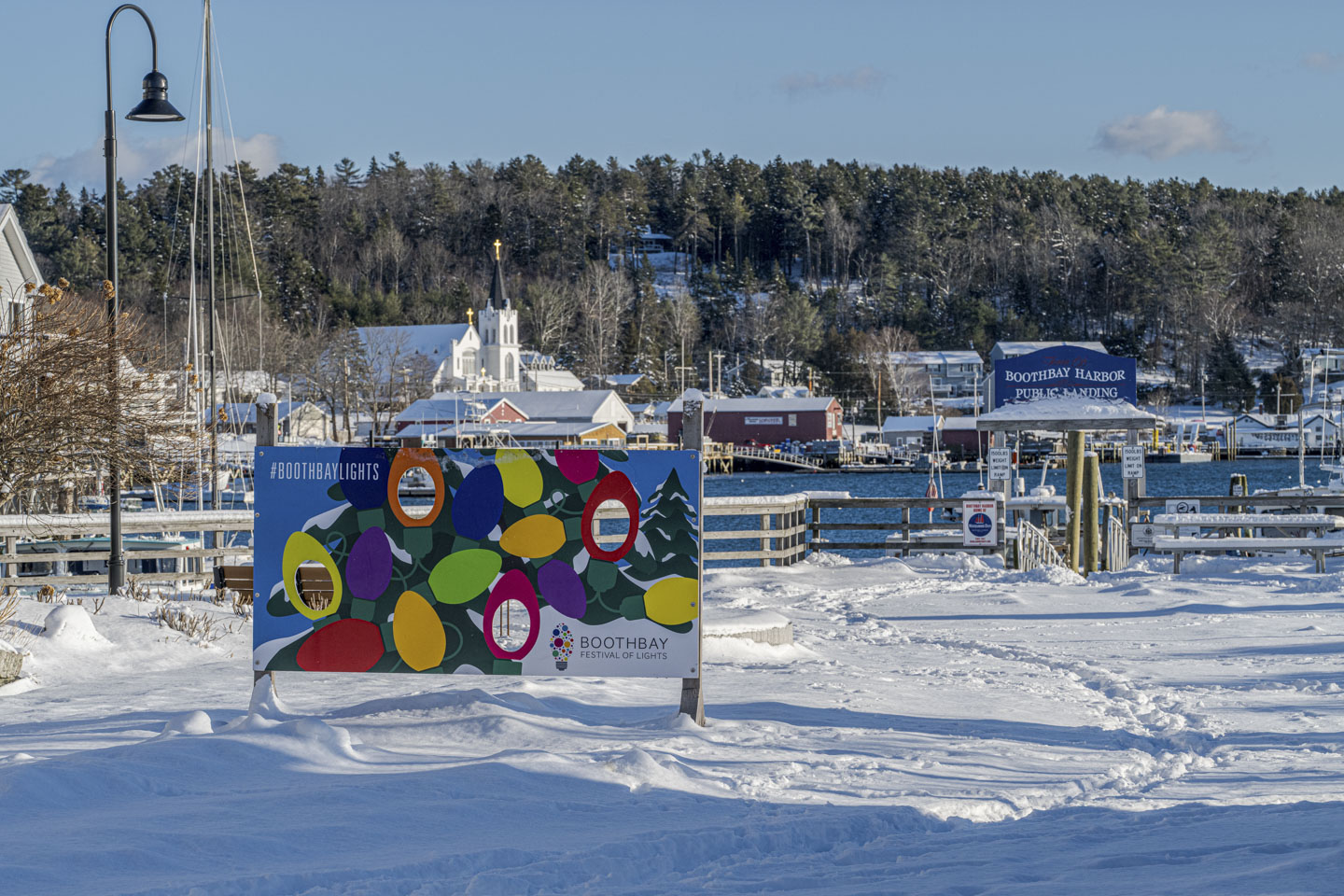 On a snowy plaza with the harbor water behind it, a colorful sign has Christmas lights painted on it.