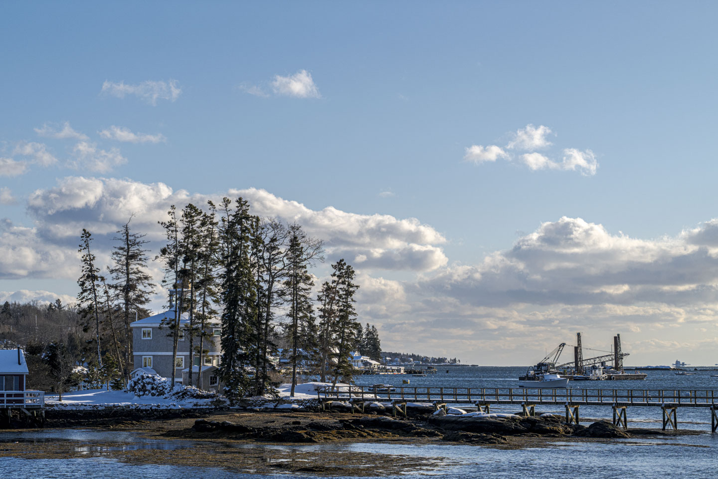 The sun is shining on a snow-covered island with tall trees and a round house, while some boats can be seen on the other side of a pier.