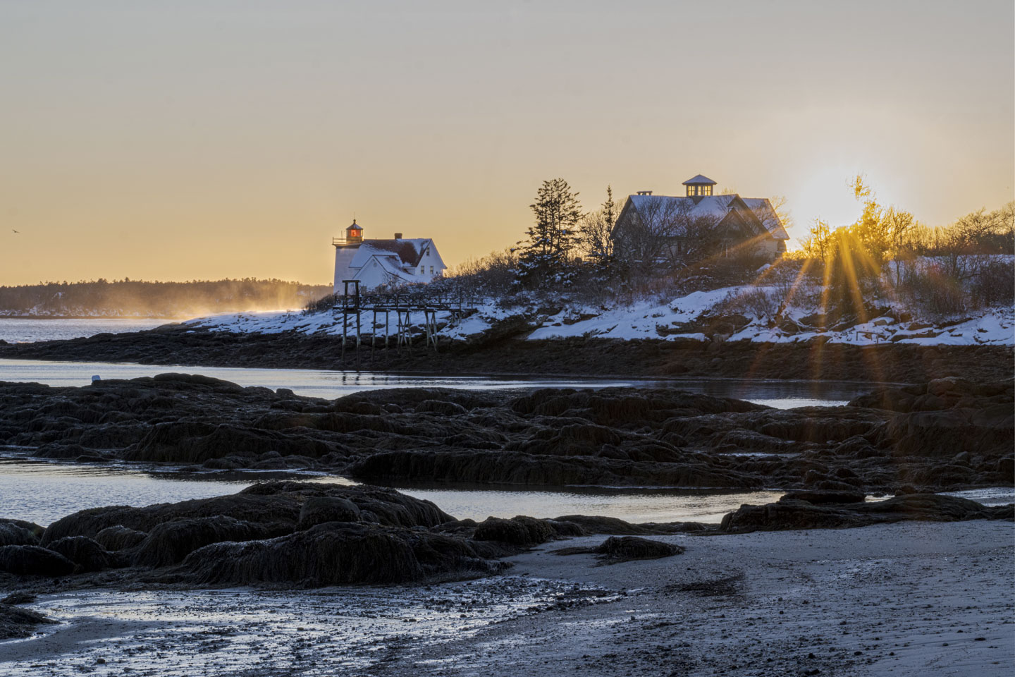 The sun is shining through trees behind a house and the lighthouse. Wind swept snow is blowing.