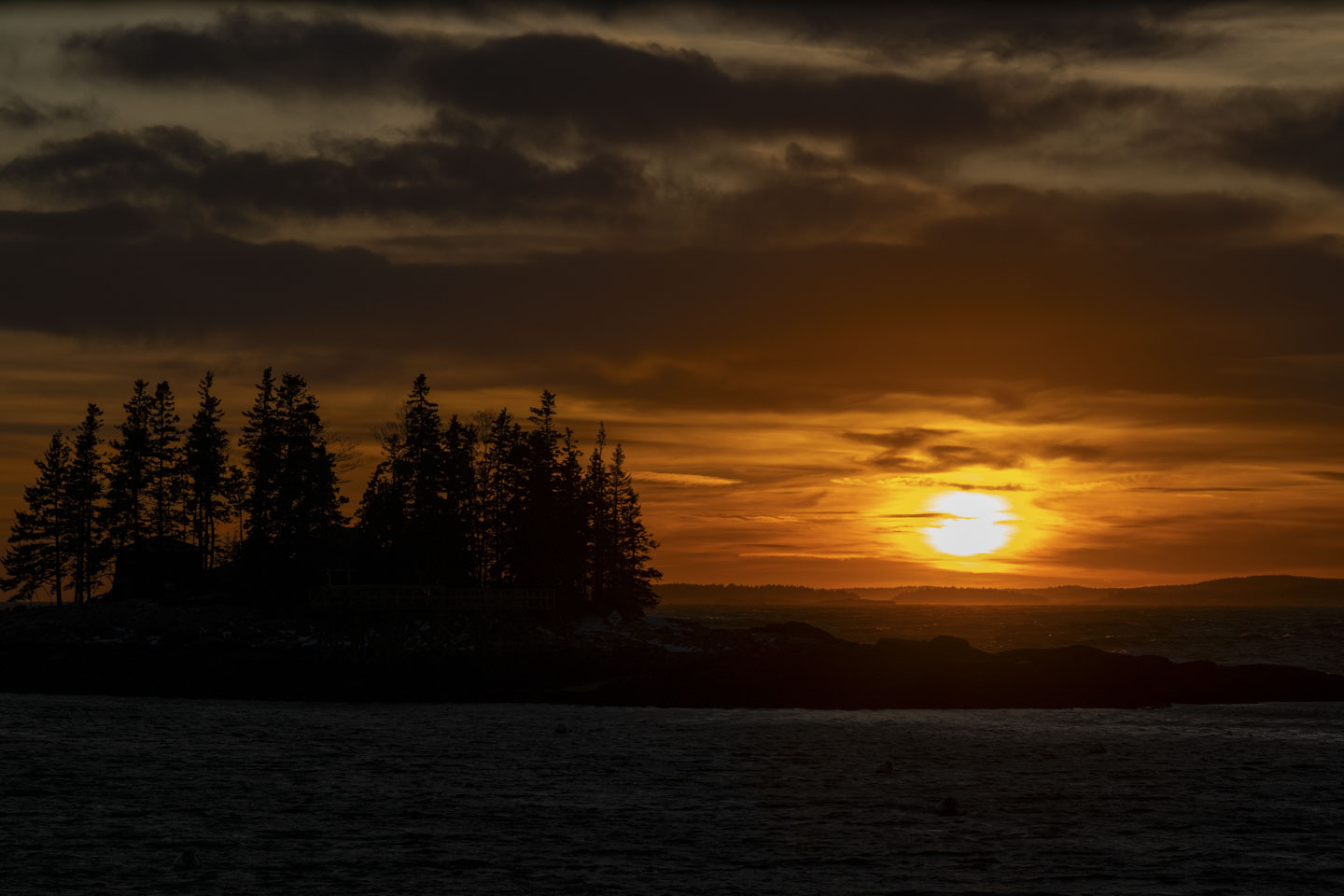 The setting sun reveals some rough water and an island with trees.