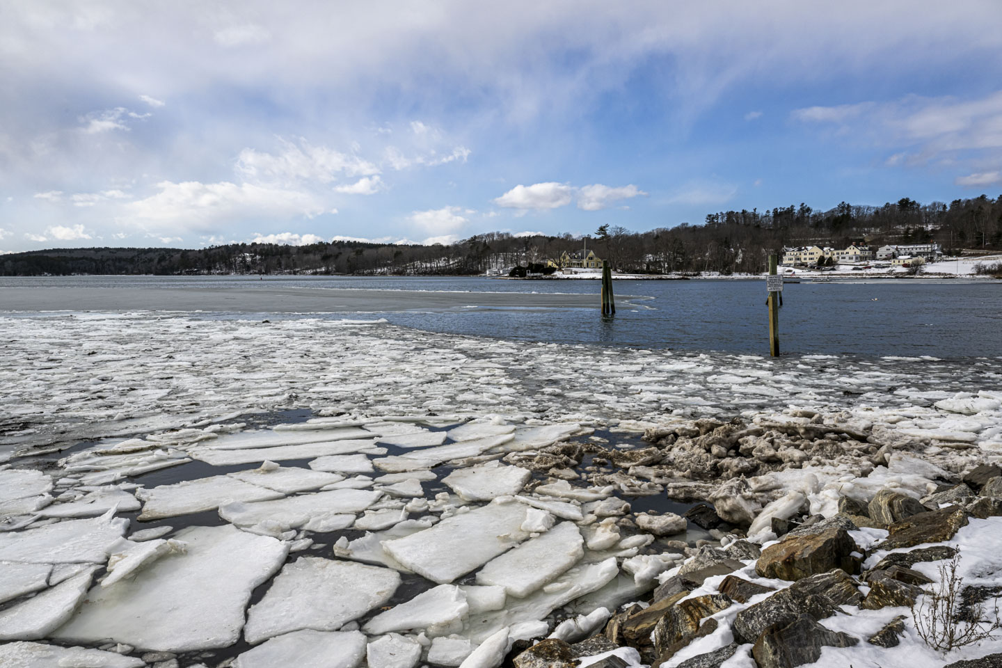 The Damariscotta River is seen with the near section covered in slabs of ice and the far section clear from ice. The sky has scattered clouds, and large houses are seen across the river.