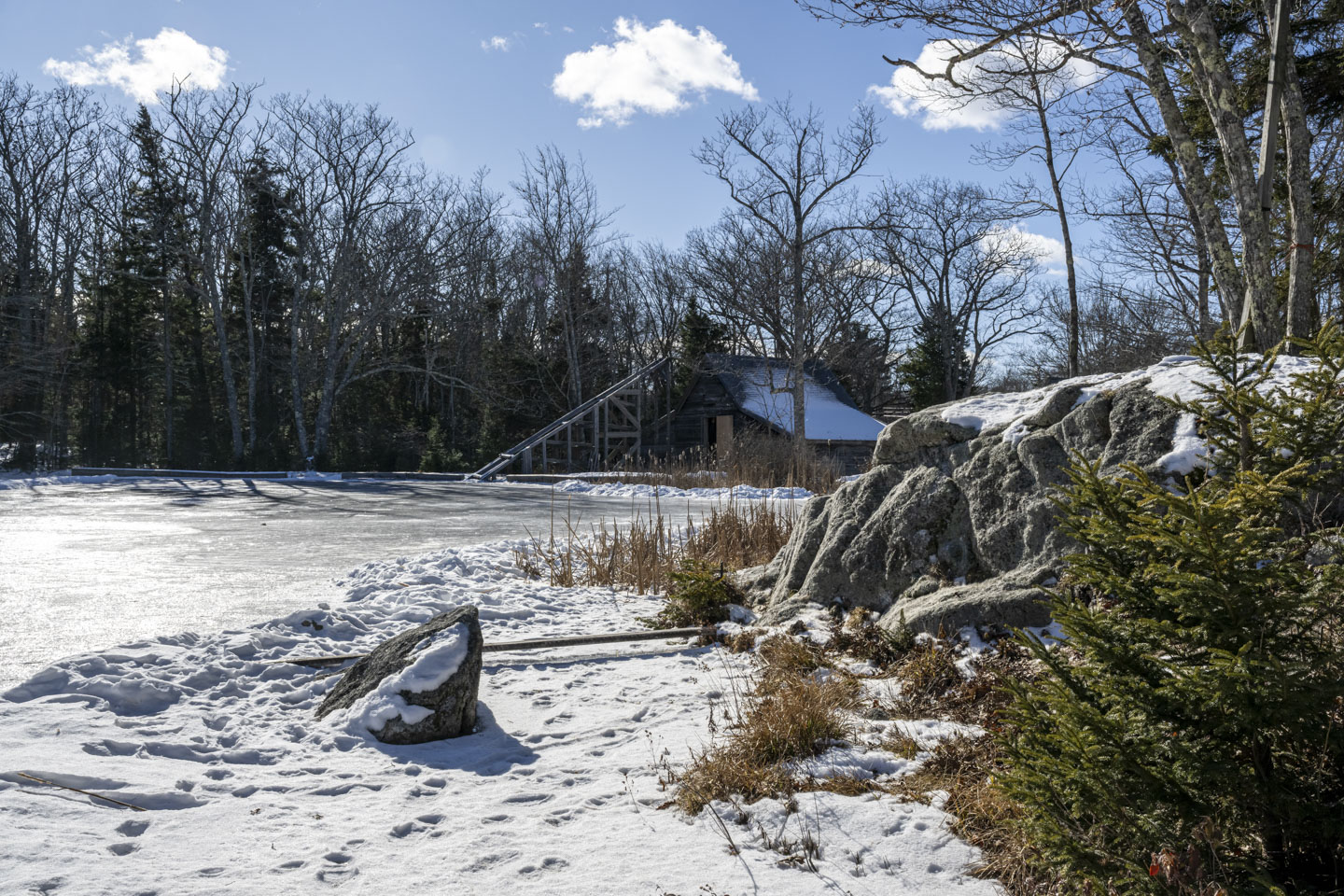 A blue sky with a few puffy clouds is above a view of a frozen pond. On the far side is a barn that has a large wooden ramp in front of it.