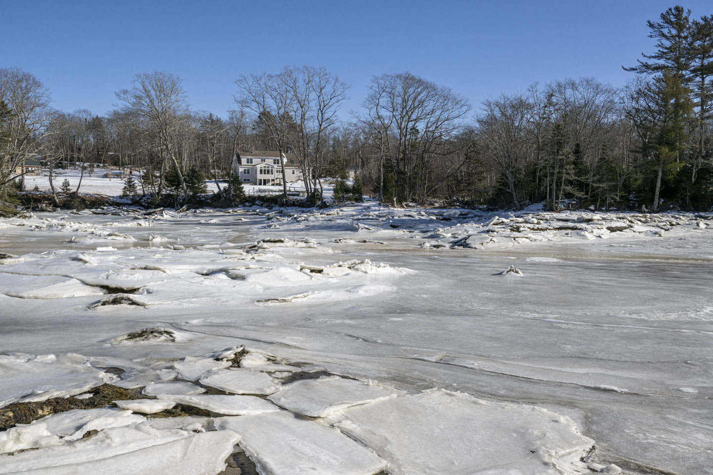 Looking back over a frozen river, you can see a large house with many windows facing the river.