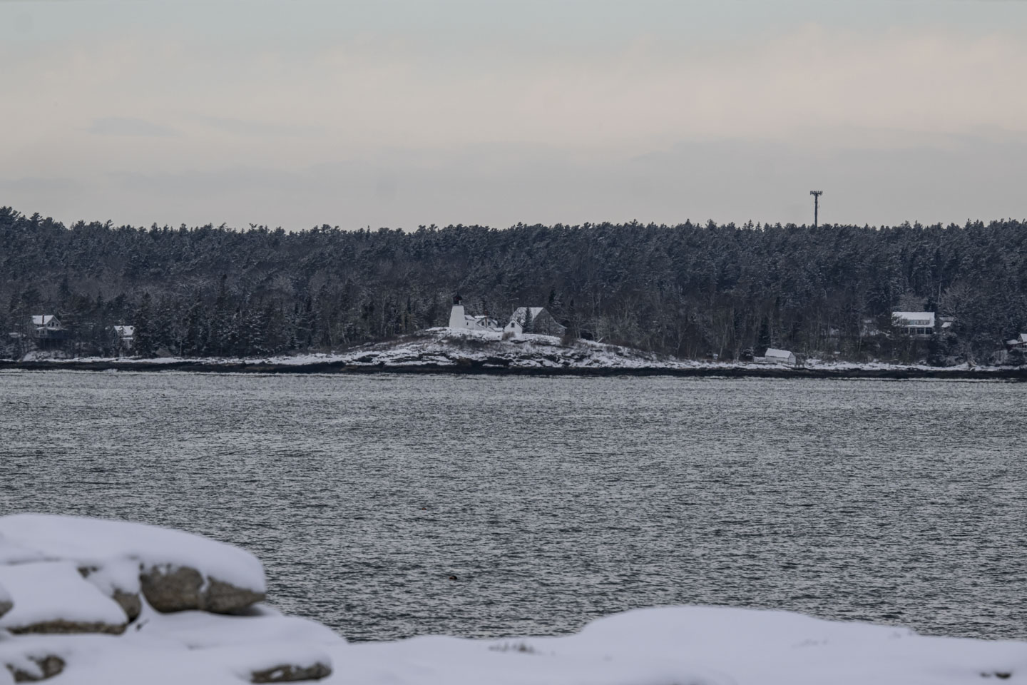 A lighthouse is on snow-covered land with trees behind it and water in front of it.