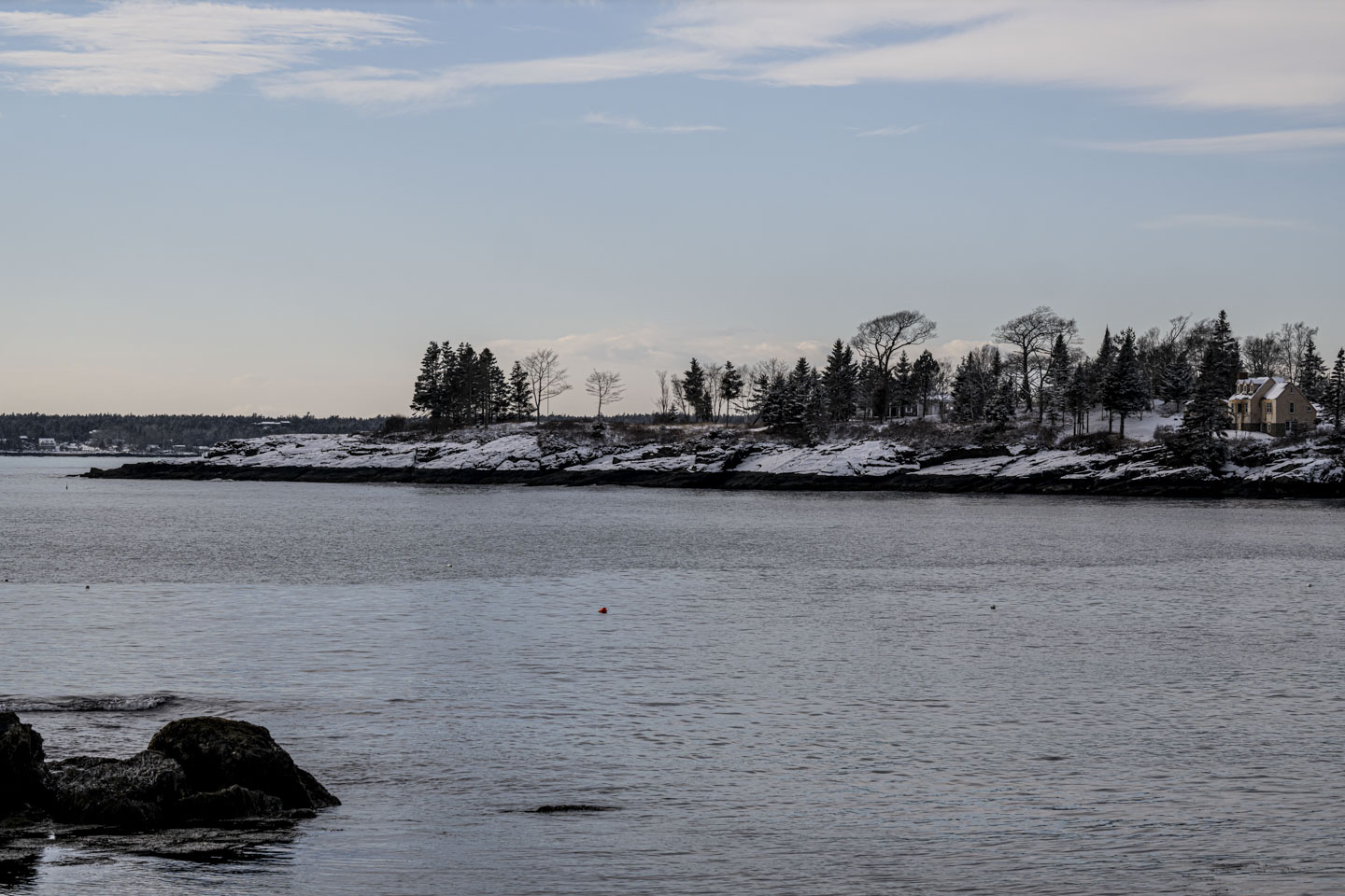 An island with snow, trees, and a couple of houses sits beneath a partly cloudy sky.