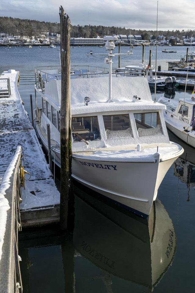 The boat Novelty is mostly covered with snow, but the dock next to is has been partially cleared.