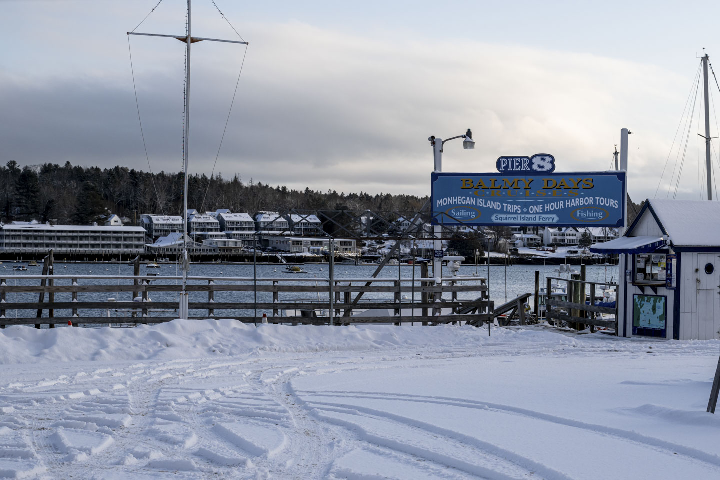 A snow-covered parking area that is next to the harbor has a large blue sign that says 'Pier 8, Balmy Days Cruises'.