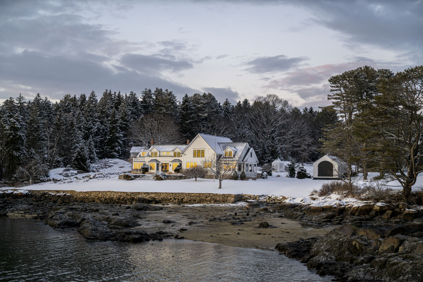 A house is on the shore of the Sheepscot River with snow covered trees behind it and a solid snow cover on the lawn. The setting sun is reflected in the windows.