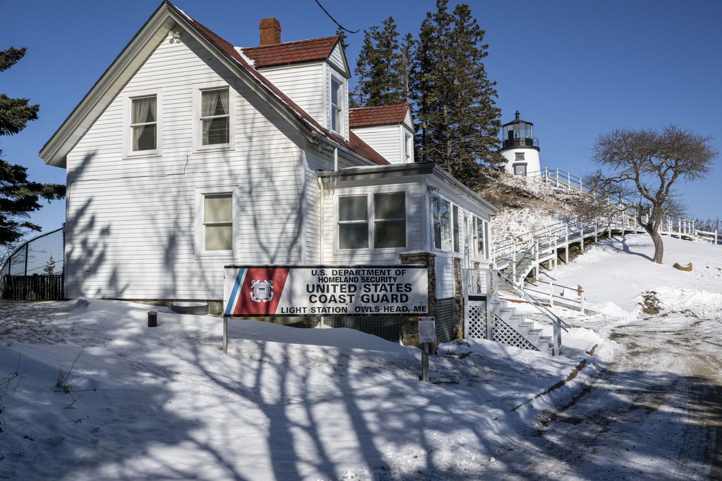 Owls Head Lighthouse is in the distance and the former light keeper cottage is closer. There is a US Coast Guard sign.