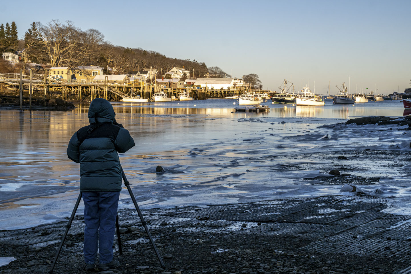 A person is standing with their back to us, and the legs of a tripod can be seen. They are looking at ice covering shallow water, with boats in the unfrozen section of the harbor. It is a sunny day.