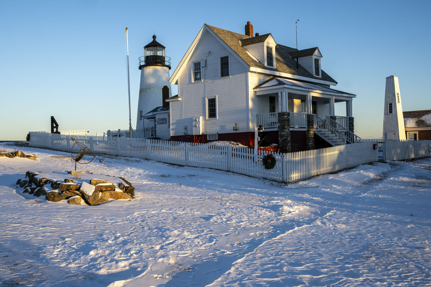 Pemaquid lighthouse is seen with snow in the yard. There is a wreath on the corner of the picket fence.