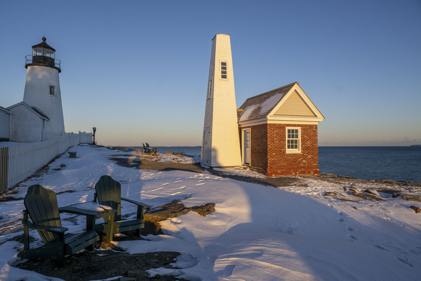 The late day sun is shining on the bell house at Pemaquid. Some Adirondack chairs sit in the snowy yard.