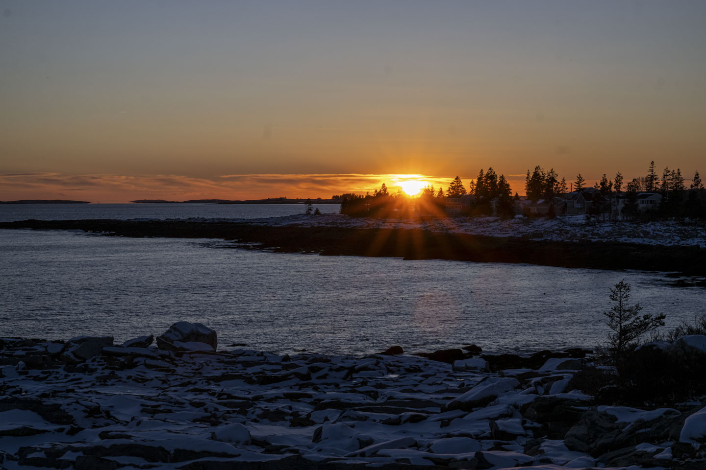 The setting sun is seen across some water and behind some trees.