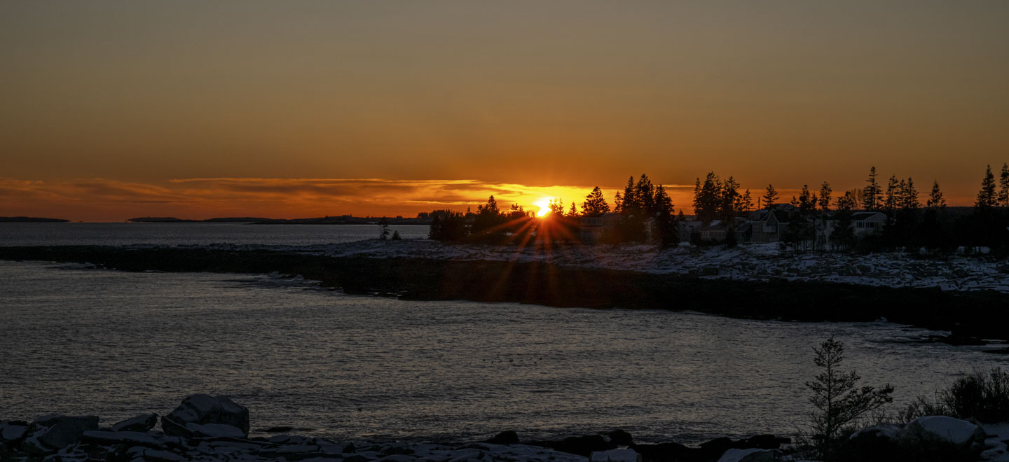 The setting sun is seen across some water and behind some trees.
