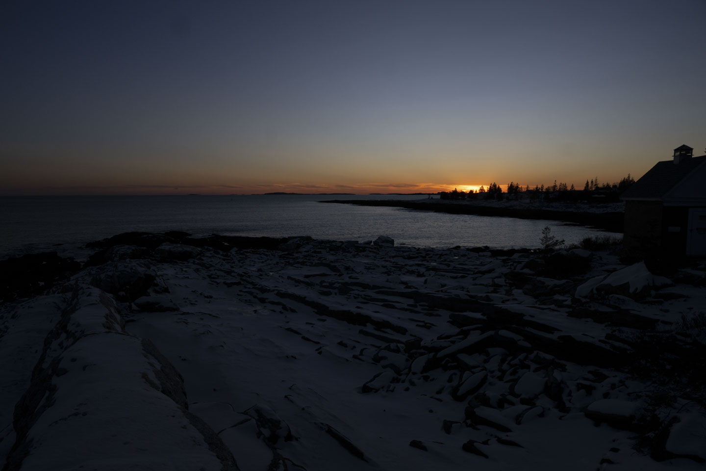 The sun is almost set in the picture. The snow-covered rocks and the water can just be seen, along with the silhouette of a house.